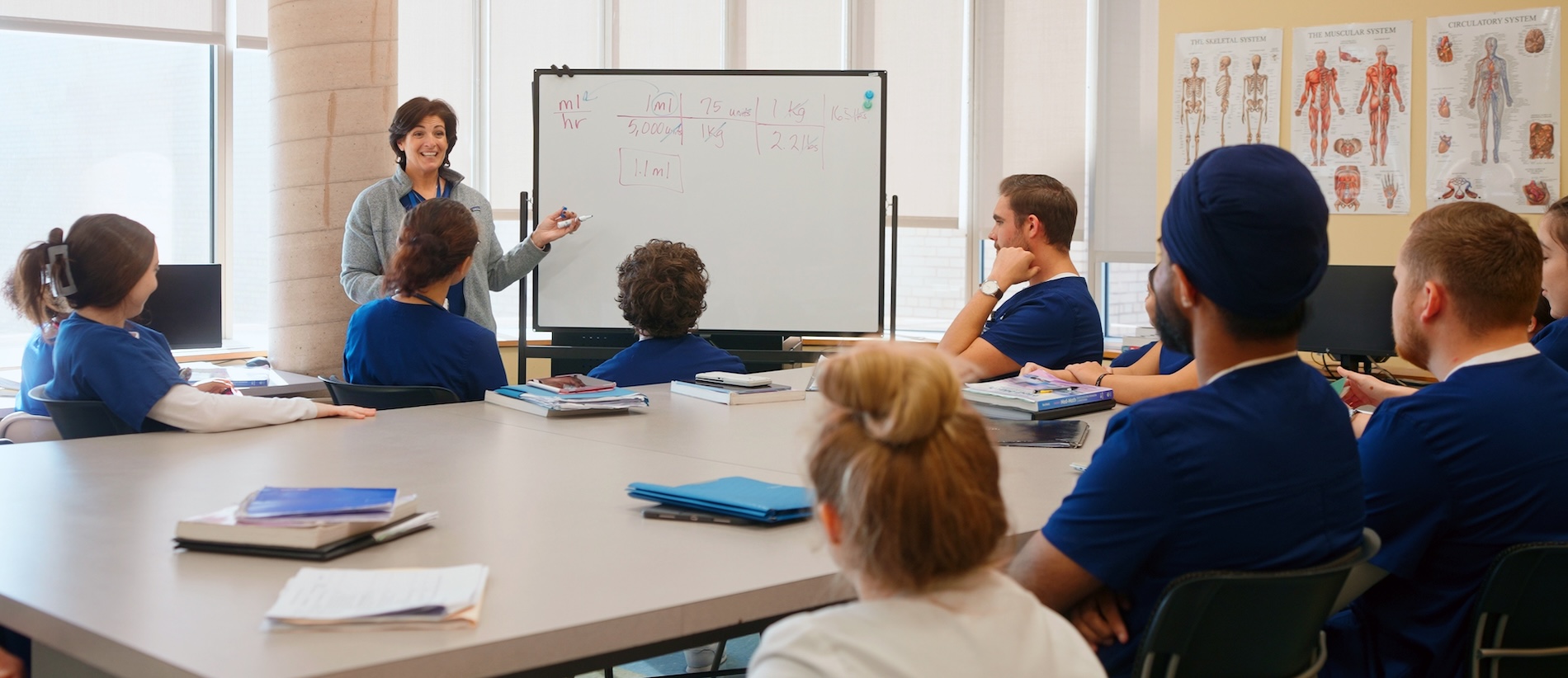 Nursing classroom with students around a class table while the professor lectures at a whiteboard