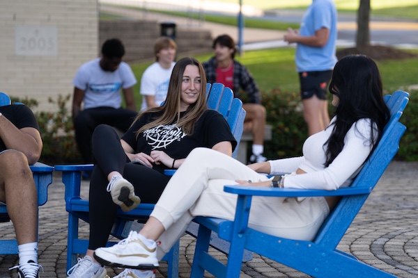 Two students sitting on blue Adirondack chairs in a plaza on campus