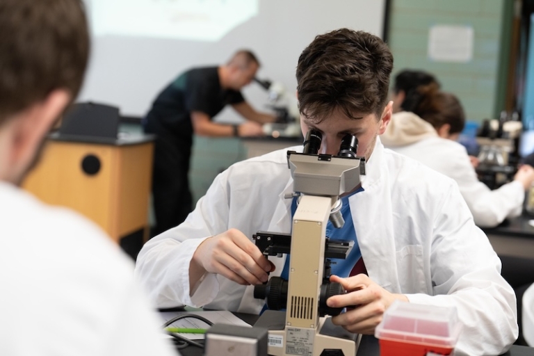 A student looks through a microscope in lab class while classmates do the same in the background