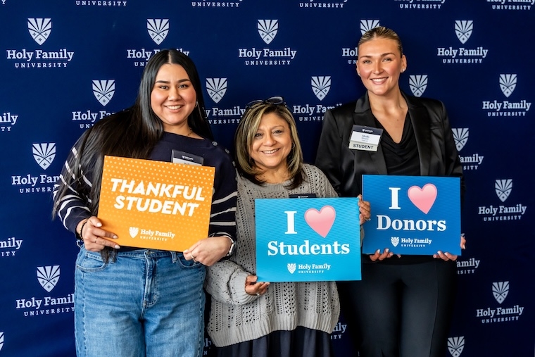 Three alum smiling while holding signs such as "Thankful Student" at an alumni event